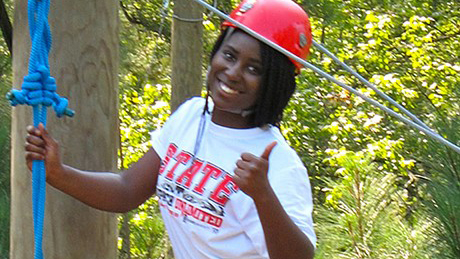 A student smiling and giving the thumbs up while having fun on a zip line.