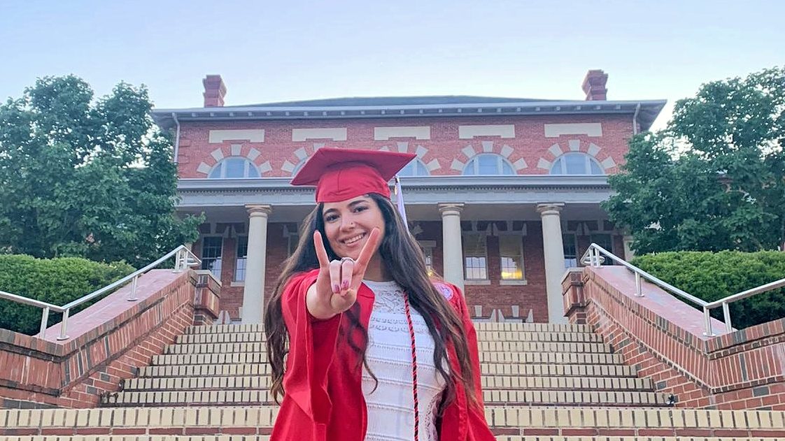 Esmira Poladova doing the wolfie hand symbol in front of a campus building in her cap and gown.