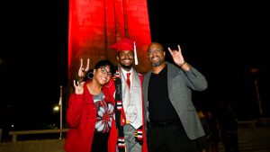 A 2021 NC State grad standing in front of the red-illuminated Memorial Belltower with his parents.