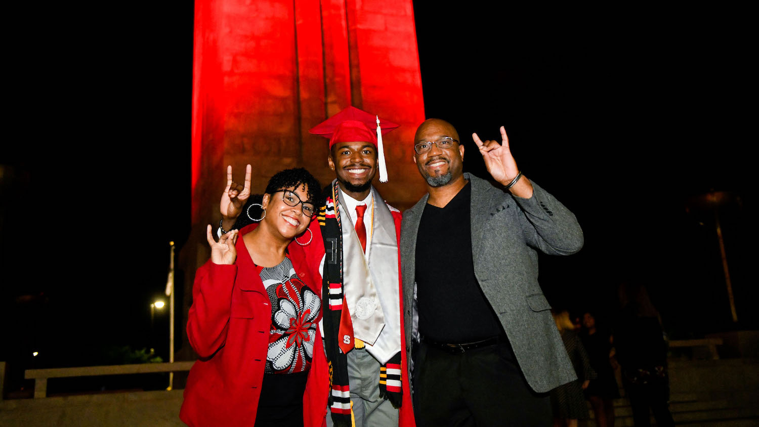 A 2021 NC&#160;State grad stands in front of the red-illuminated Memorial Belltower with his parents