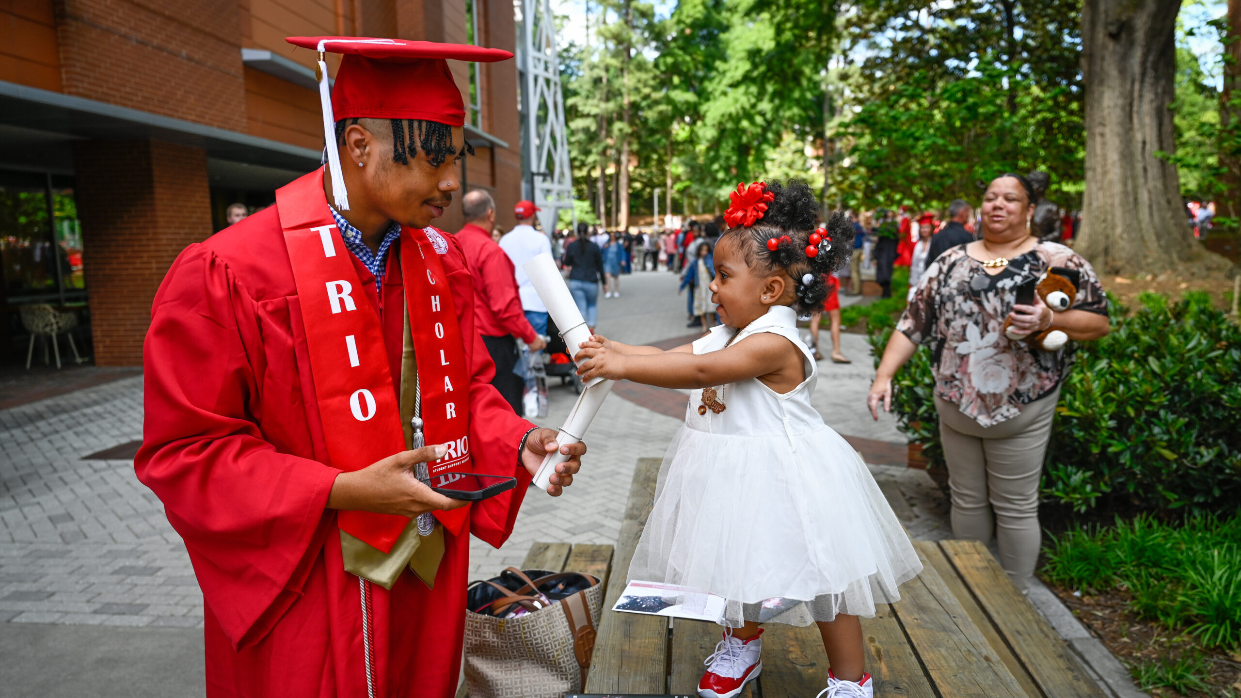 a TRIO graduate stands in his cap and gown with a small child